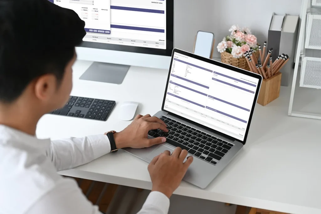 A person working on a laptop and desktop monitor displaying what appears to be a Ubersmith billing system interface, customer management software for hosting providers.