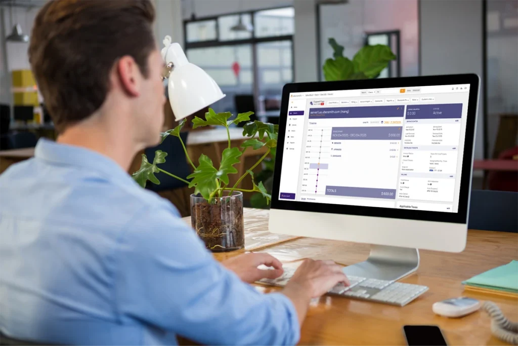 A person in a blue shirt working at a desktop computer displaying the Ubersmith billing platform, showing a client account summary with service details, billing period, and invoice totals, data center billing software.