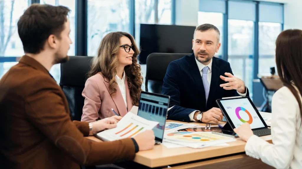 Four business professionals in a meeting room reviewing charts and data on printed reports and a tablet, engaged in a discussion about operational metrics, usage-based billing software.