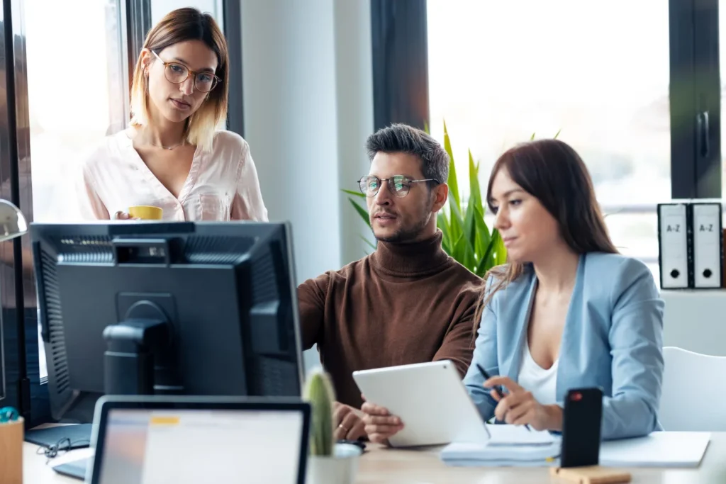 Three business professionals reviewing operational data together on a desktop monitor and tablet in an office setting, network operations management software.