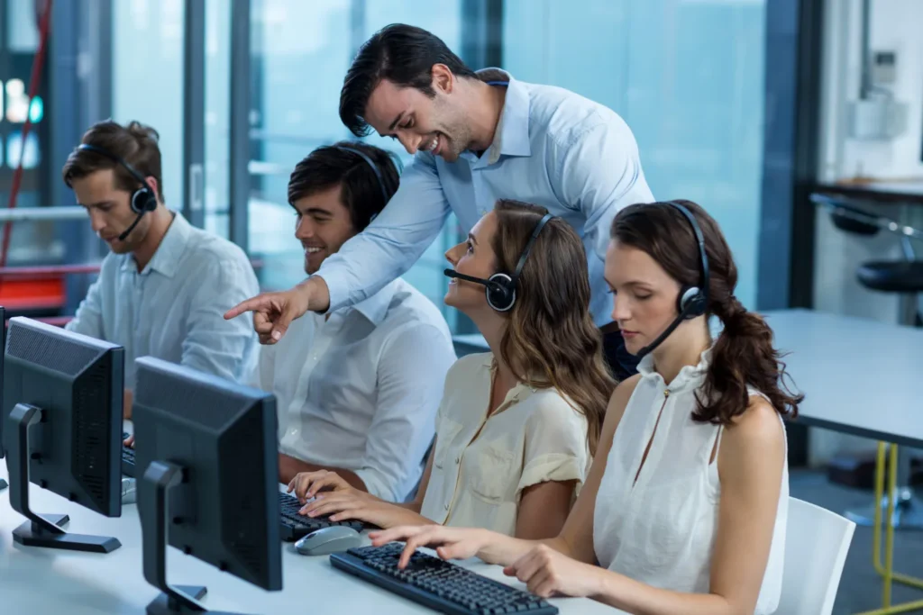A team of telecom support agents wearing headsets at their workstations, with a supervisor pointing at a monitor during an active operations session, automated telecom billing system in USA.