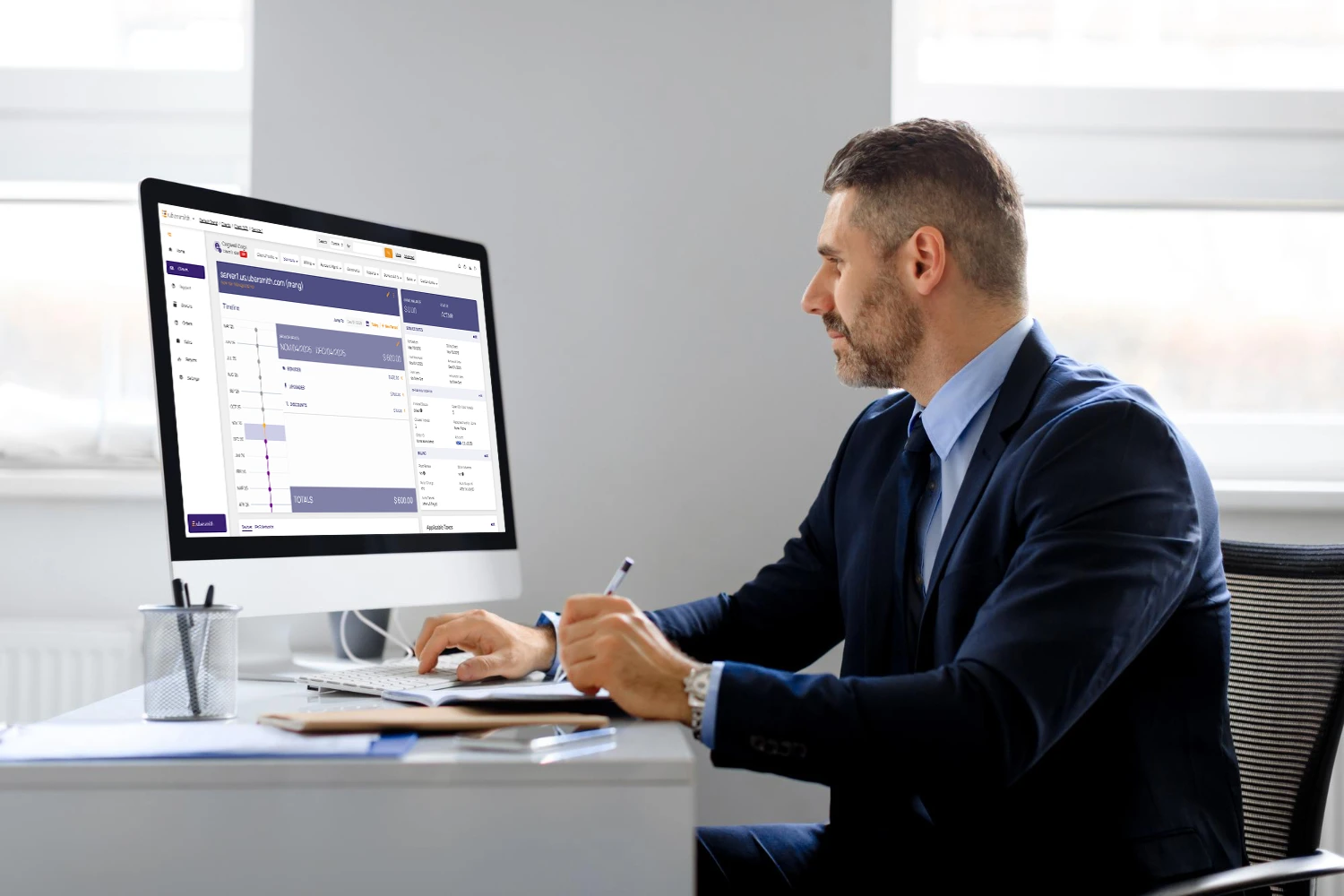 A man in a business suit sitting at a desk, reviewing data on a large desktop monitor displaying a Ubersmith software dashboard interface, AI infrastructure management platform for ISPs US.